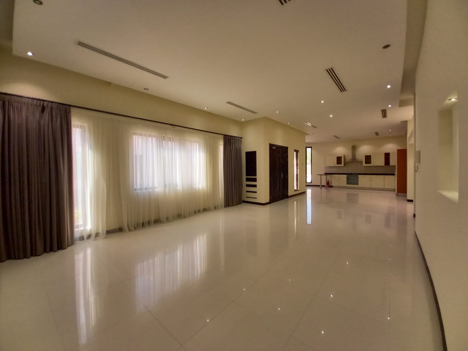 Spacious, empty living area with glossy tile flooring, large windows with curtains, and recessed ceiling lights, leading to an open kitchen at the far end.