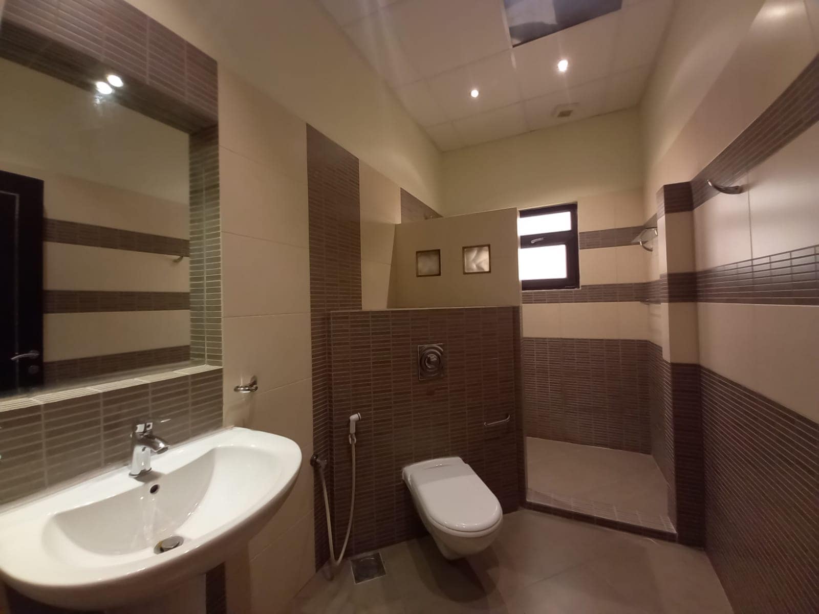 A modern bathroom with a white sink, a wall-mounted toilet, and a shower area separated by a tiled partition. The walls are tiled in brown and beige.