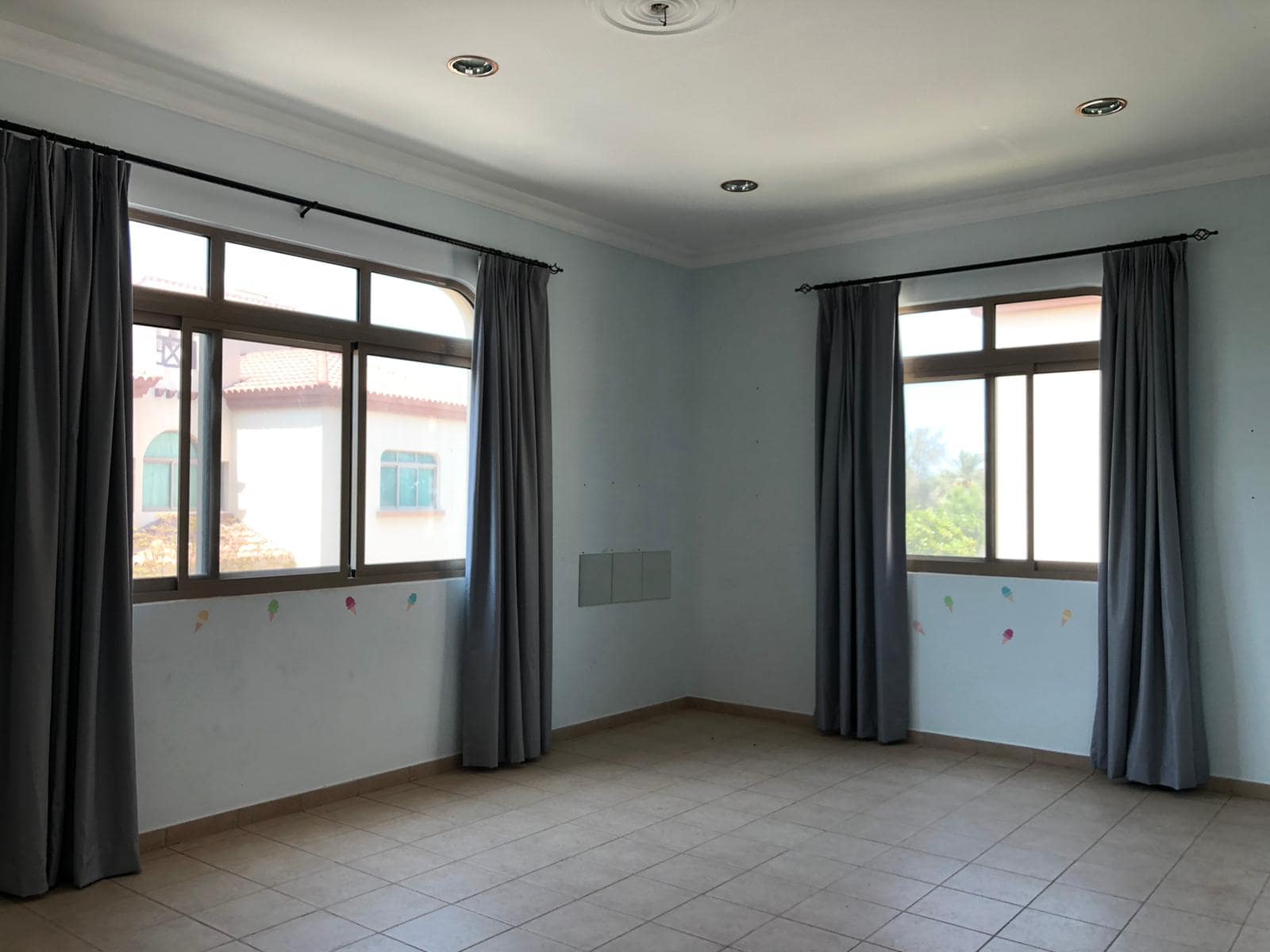 Empty room with two large windows covered by gray curtains. The walls are light blue with faint marks and the floor is tiled. Ceiling lights are also visible.