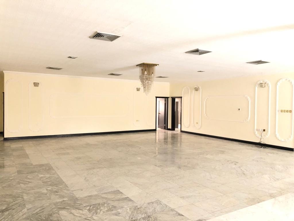 A spacious, empty room with marble flooring, light-colored walls, modern light fixtures, and a crystal chandelier hanging from the ceiling. Several doorways and vents are also visible.