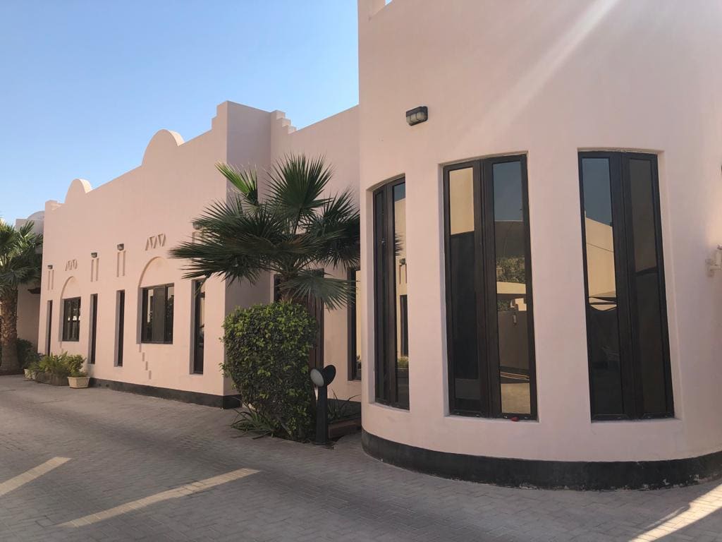 A modern, single-story building with a light exterior, large dark-tinted windows, palm trees, and paved walkway in front.