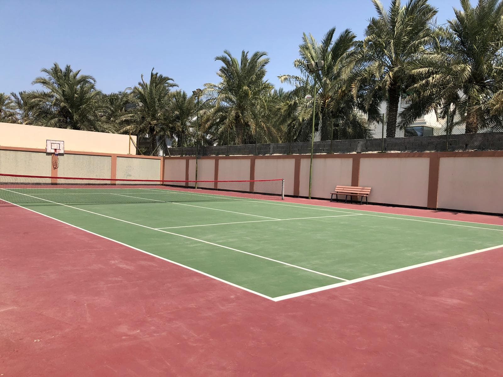 Outdoor tennis court with green playing area surrounded by red surface, set against a backdrop of palm trees and a clear sky.