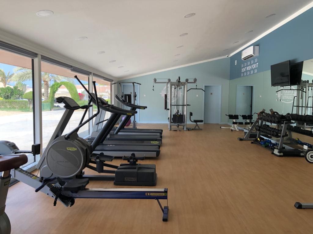 A spacious, well-lit gym with treadmills, elliptical machines, weight machines, free weights, and a TV on the wall. Large windows offer a view of an outdoor area.