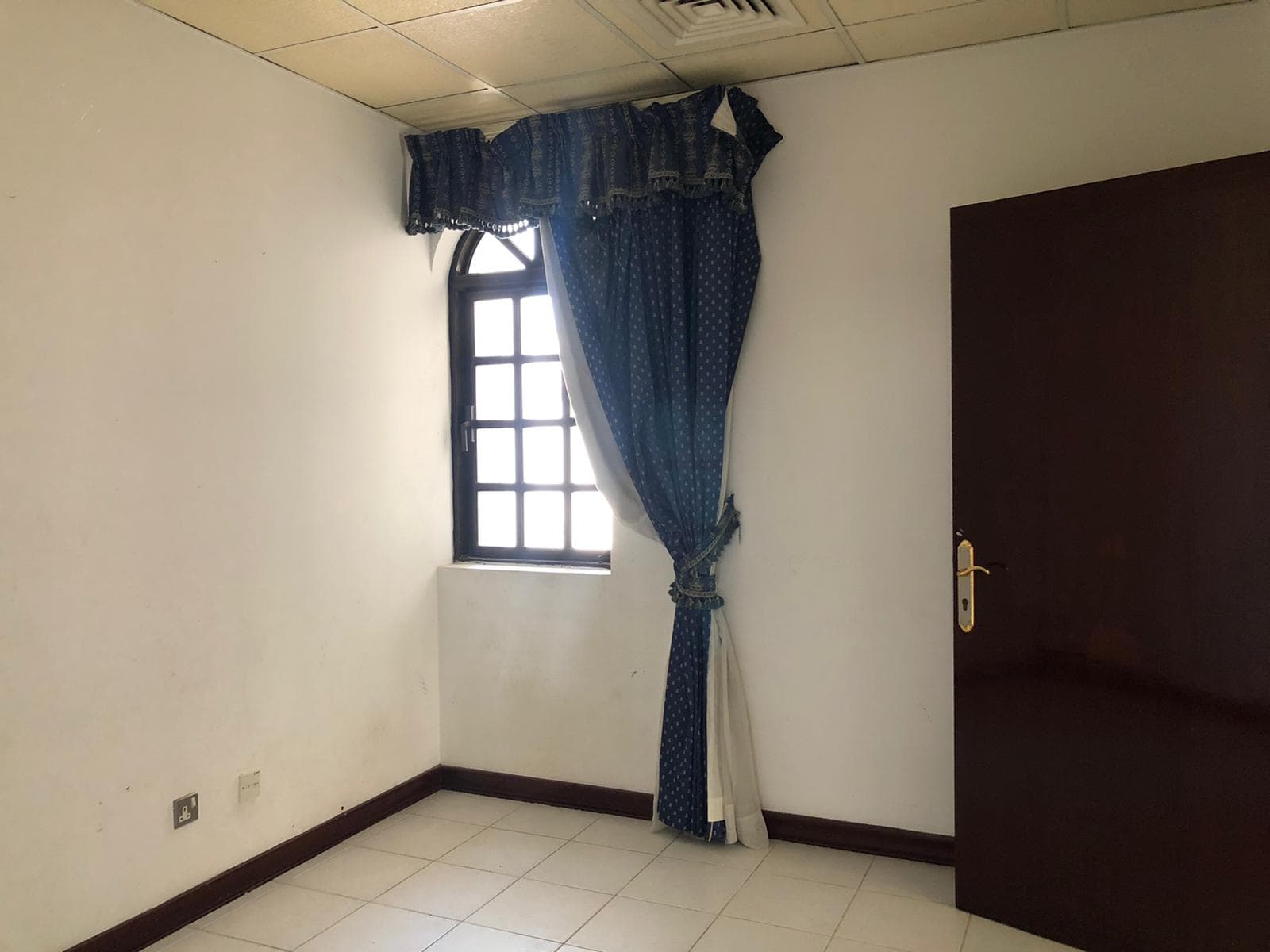 An empty room with a small window covered by blue curtains tied back, a white tiled floor, white walls, and a brown wooden door partially open.