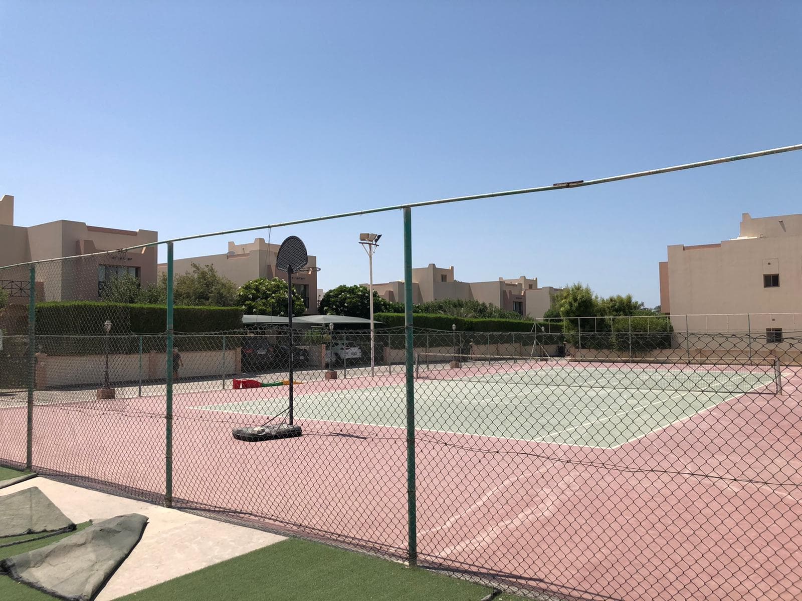 A fenced outdoor tennis court in a residential area with several low-rise buildings and greenery in the background under a clear blue sky.