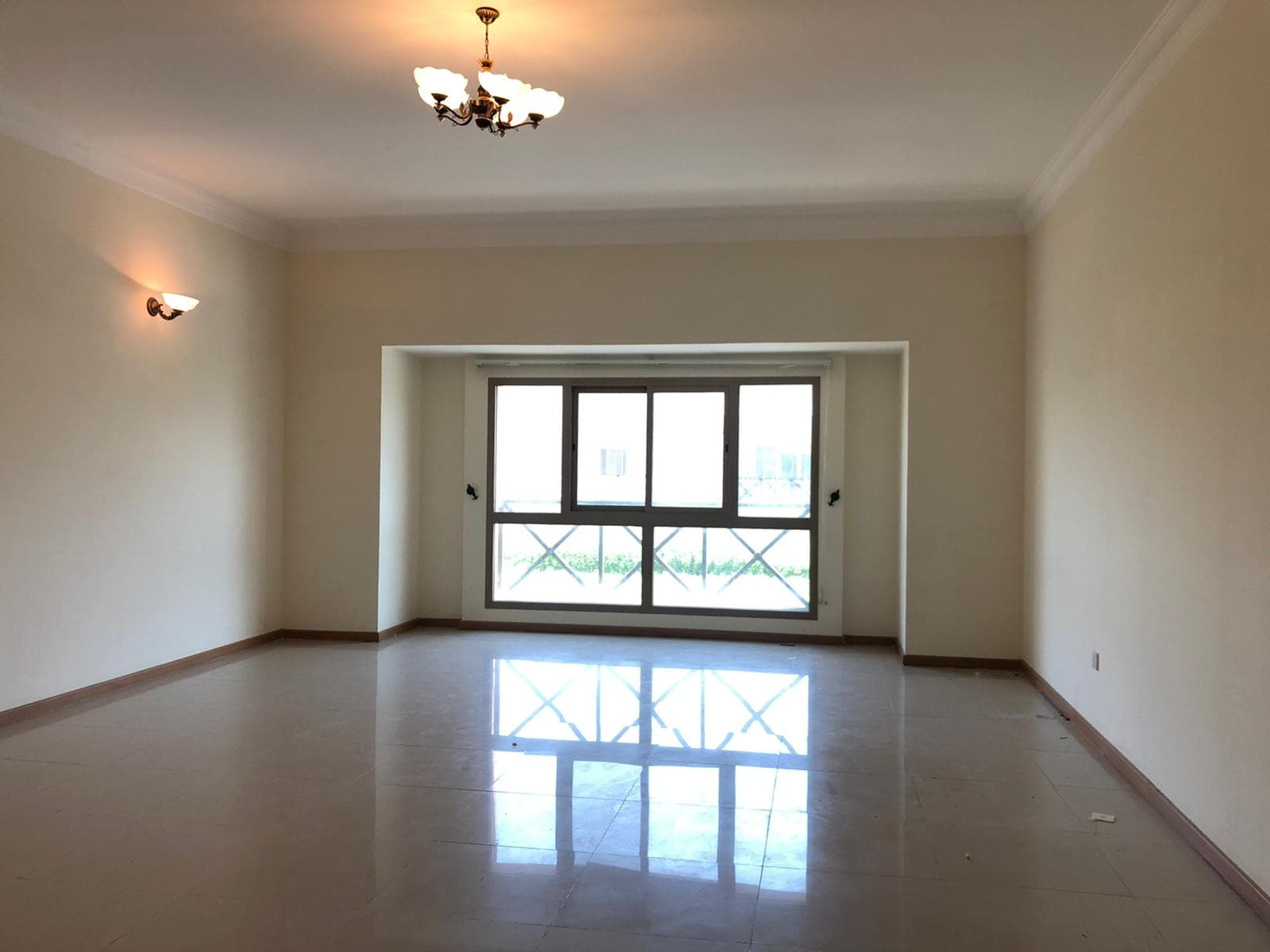 An empty room with cream-colored walls, a large window, and a chandelier on the ceiling. The floor is tiled and there is a wall-mounted light fixture on the left wall.