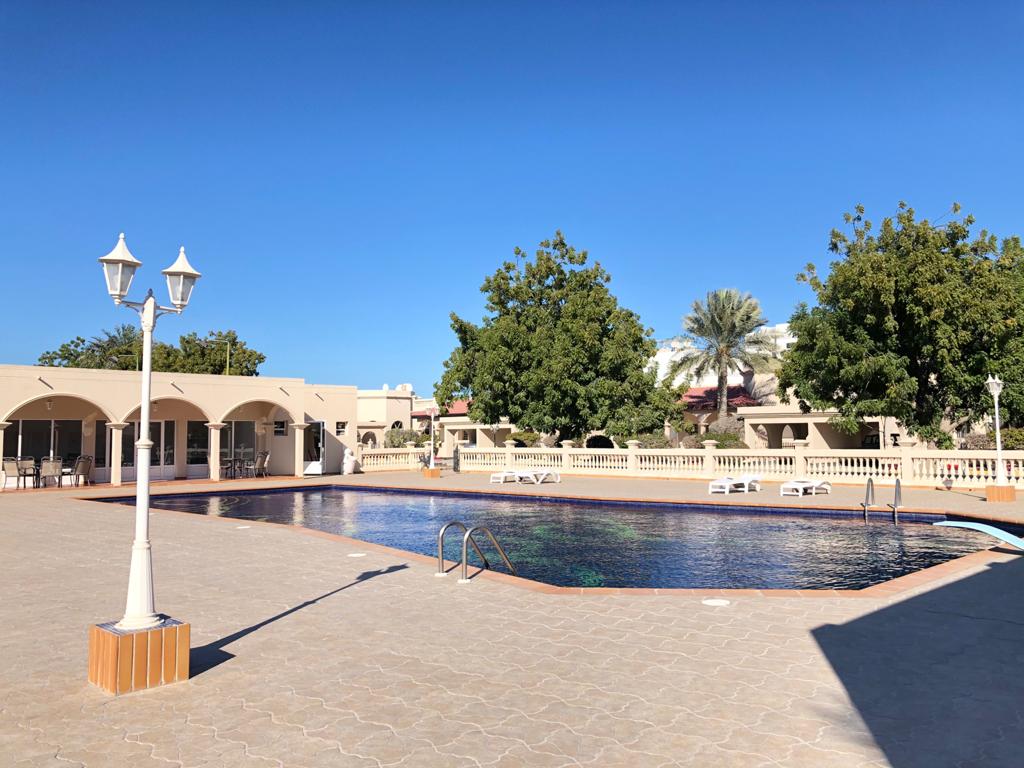 Outdoor swimming pool with clear water, surrounded by a tiled deck and lamp posts. There are several lounge chairs and green trees in the background under a clear blue sky.