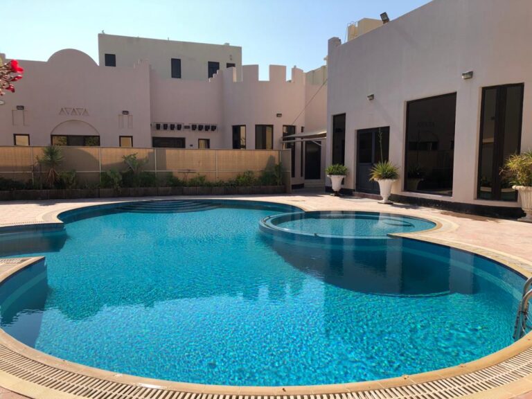 A residential backyard featuring a large, clear blue swimming pool surrounded by modern white buildings and potted plants.