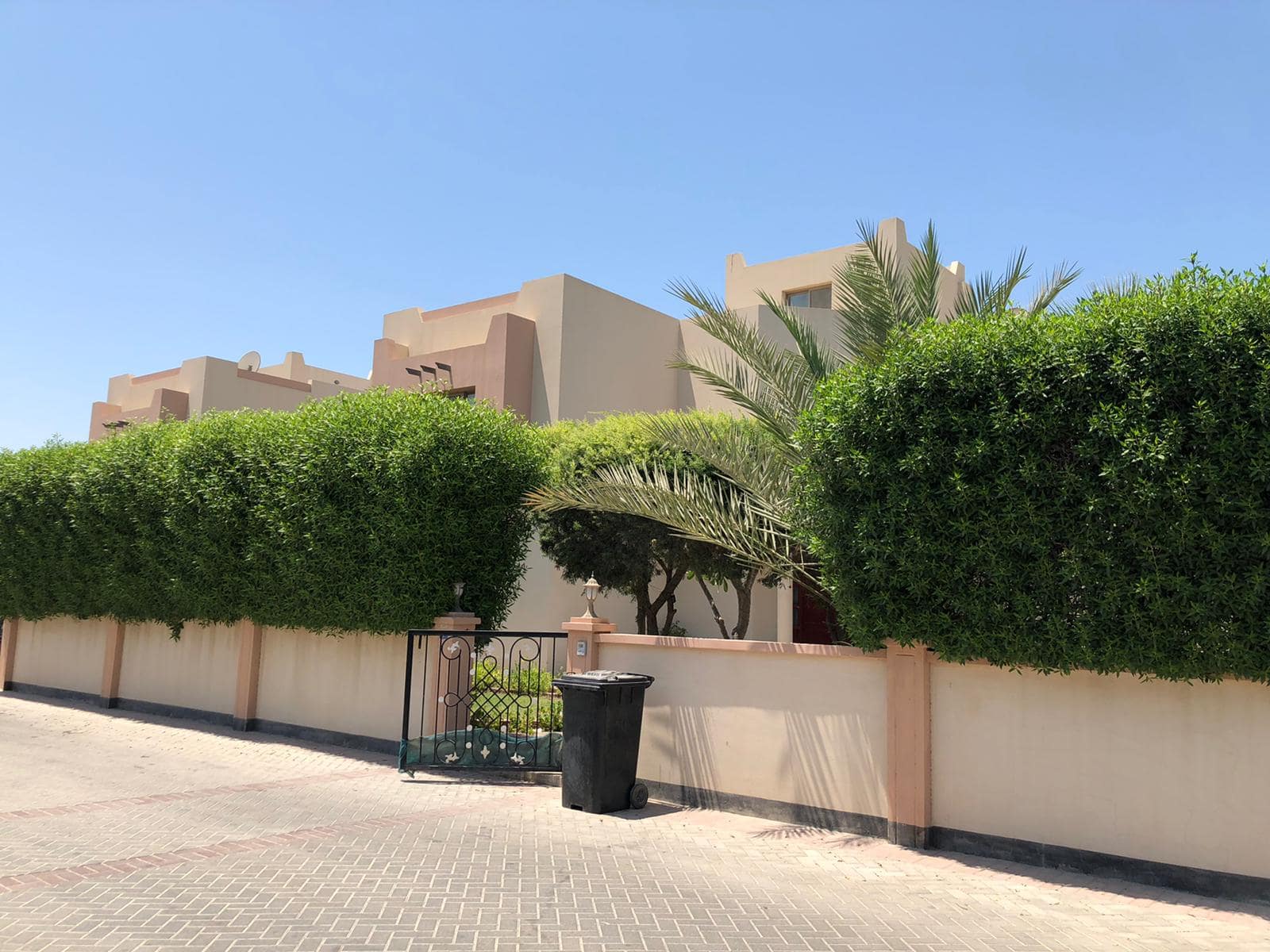 A beige building with a flat roof is partially obscured by a tall, well-manicured hedge and palm tree. A black trash bin stands next to a gated entrance, and a clear sky is visible in the background.