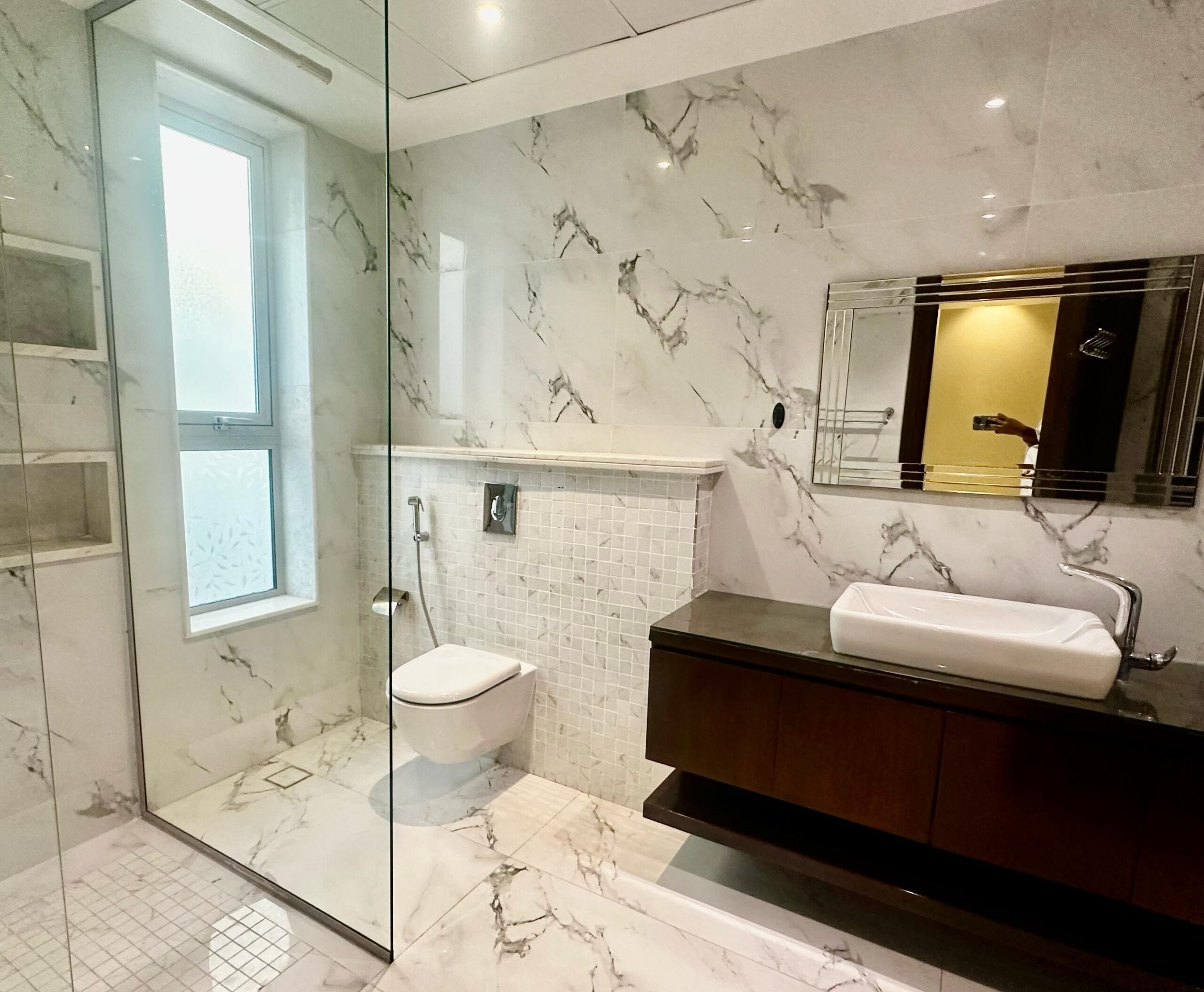 A modern bathroom with a glass-enclosed shower, wall-mounted toilet, large mirror, vessel sink, and marble tile walls and floors.