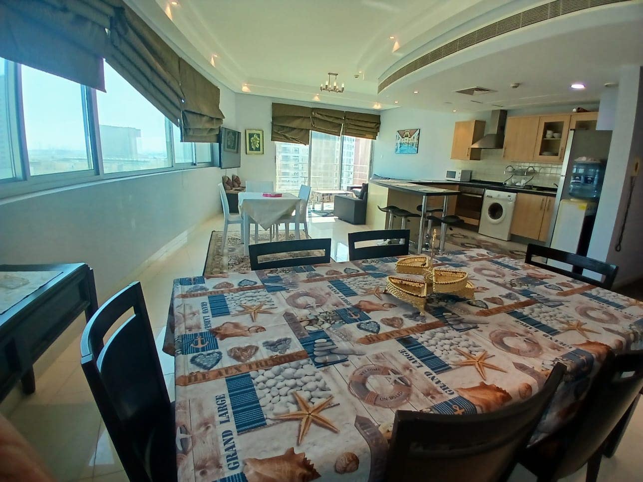 A modern kitchen and dining area with ocean-themed tablecloth, large windows, and an open-plan layout. A small dining table and chairs are visible in the background near the windows.