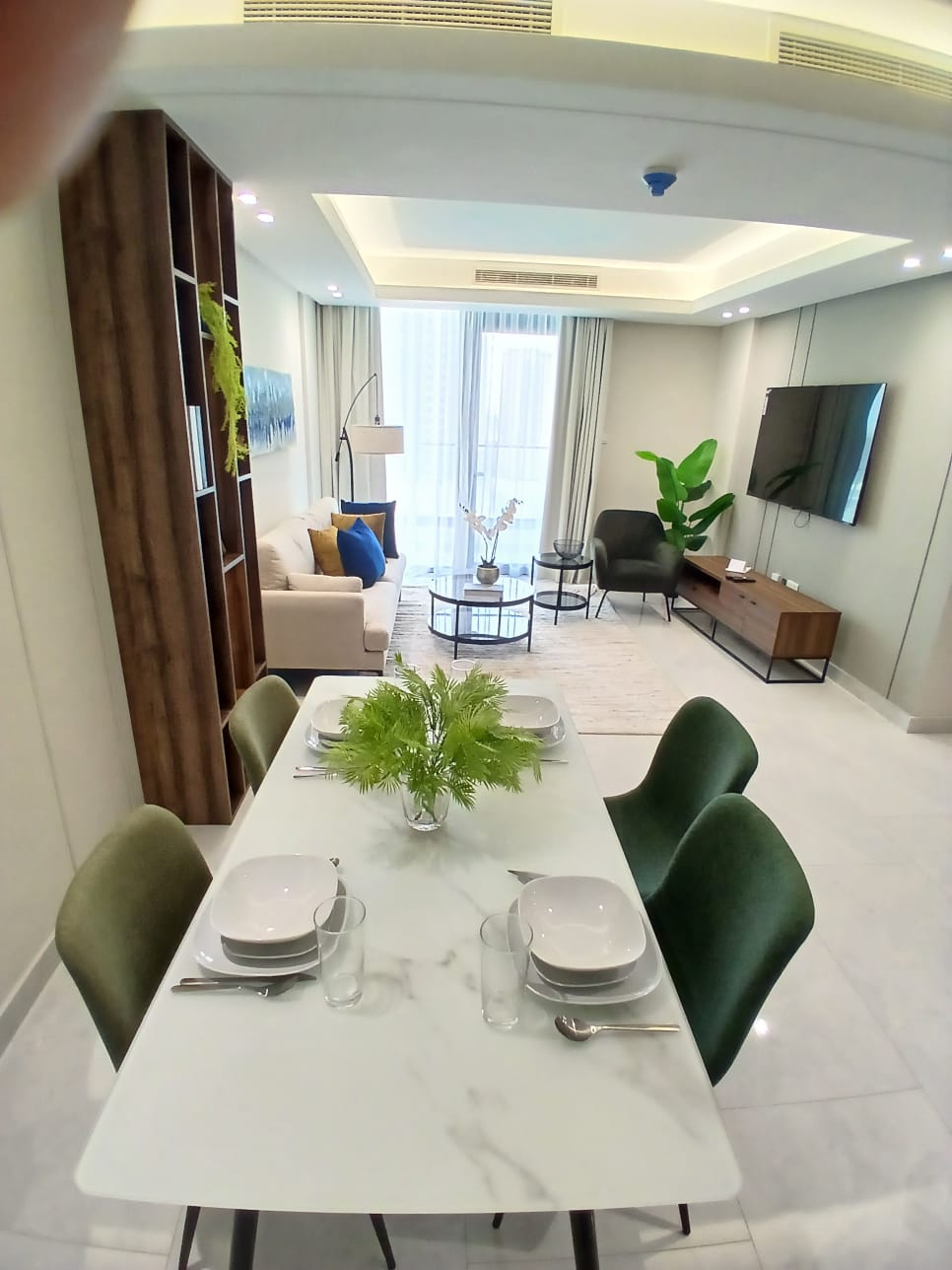 A modern living and dining area with a white dining table set for four, a cozy sofa, armchairs, a TV on the wall, a coffee table, and a large plant. Sliding glass doors lead to an outdoor space.