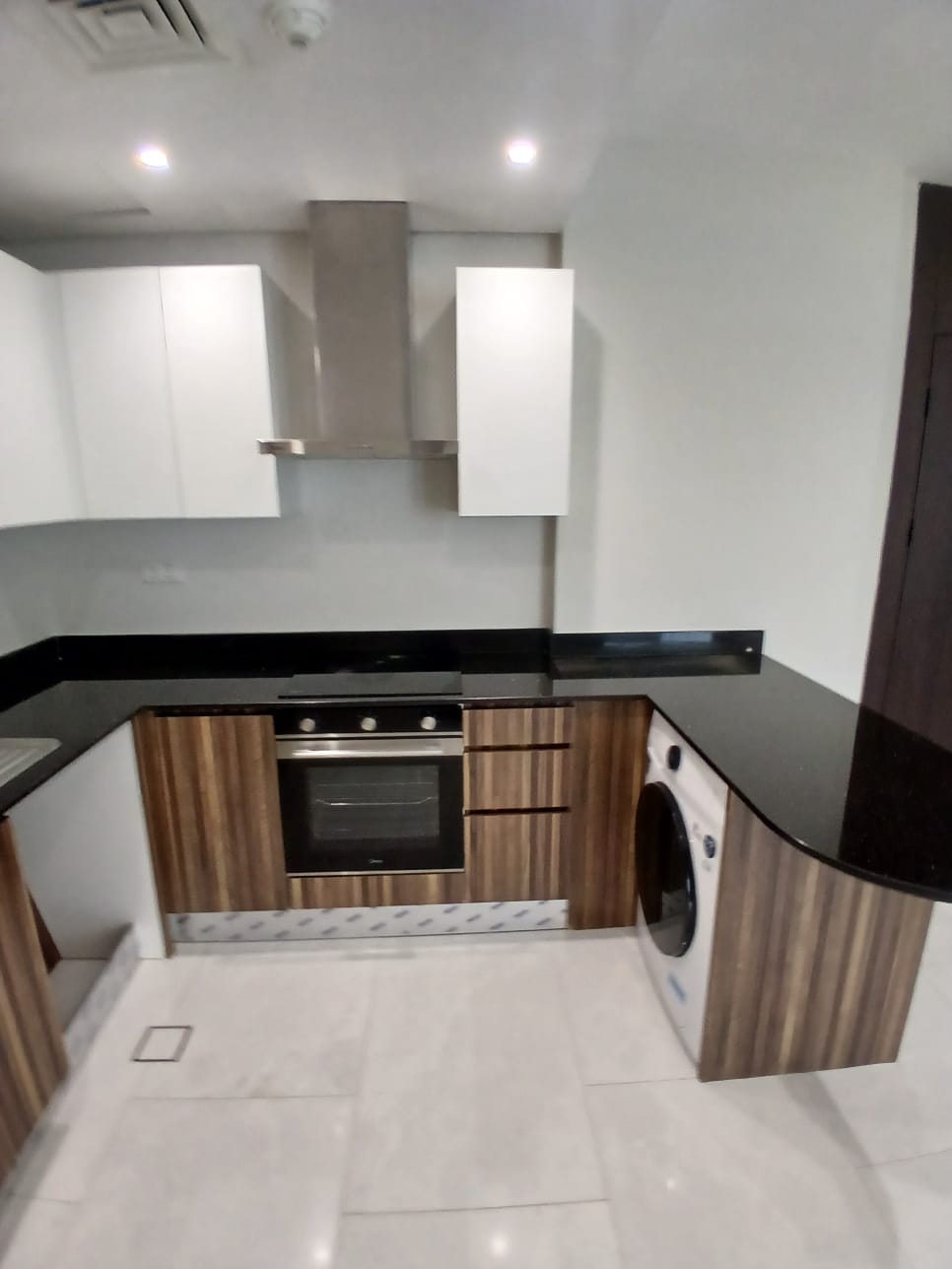 A modern kitchen with wood cabinetry, black countertops, an oven, a stovetop with an exhaust hood, and a washing machine. The floor is tiled in a light color.