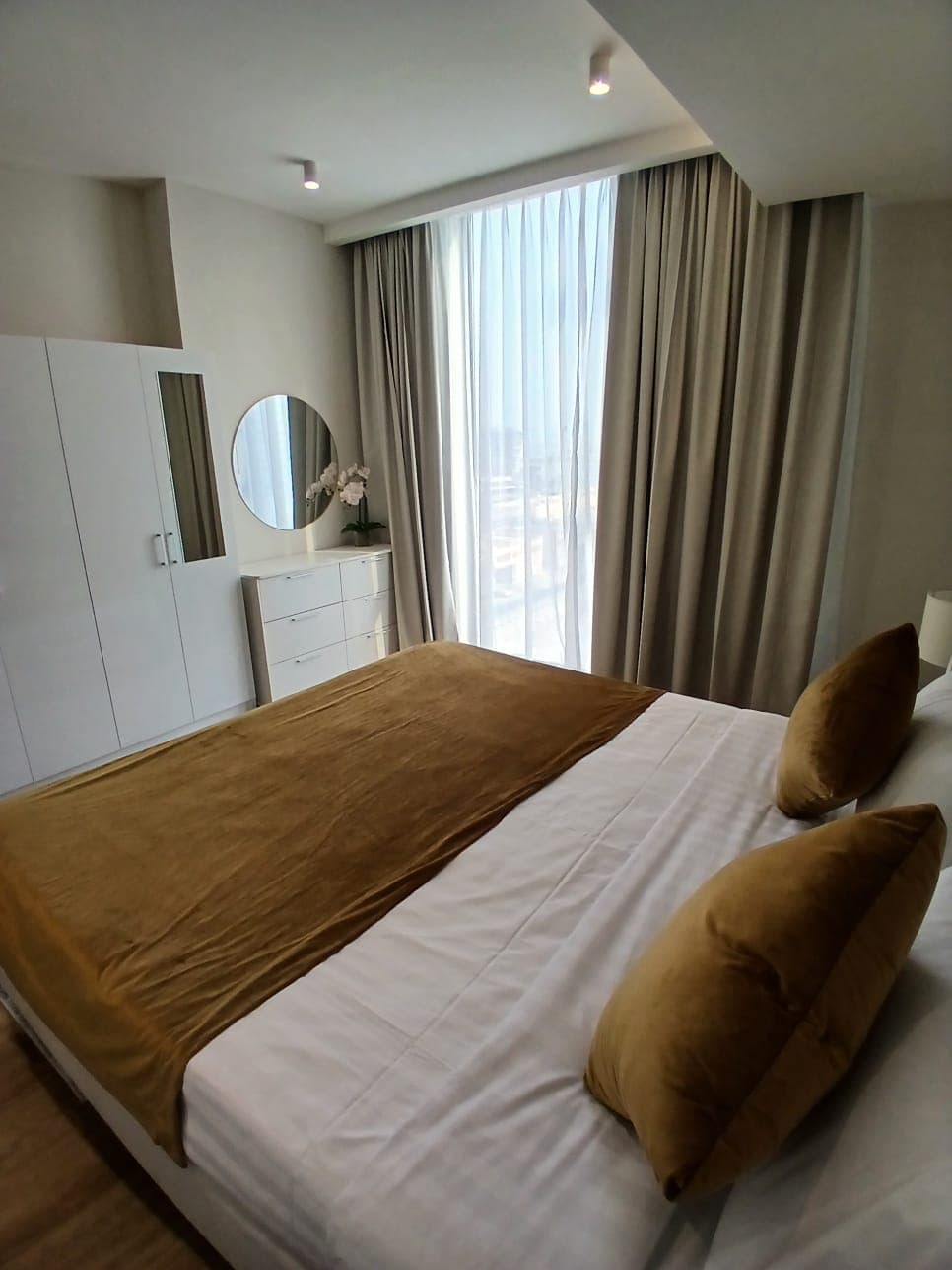 A modern bedroom with a made bed featuring brown pillows and a brown bedspread. It has curtains, a round mirror above a white dresser, and a white wardrobe. Light filters through a large window.