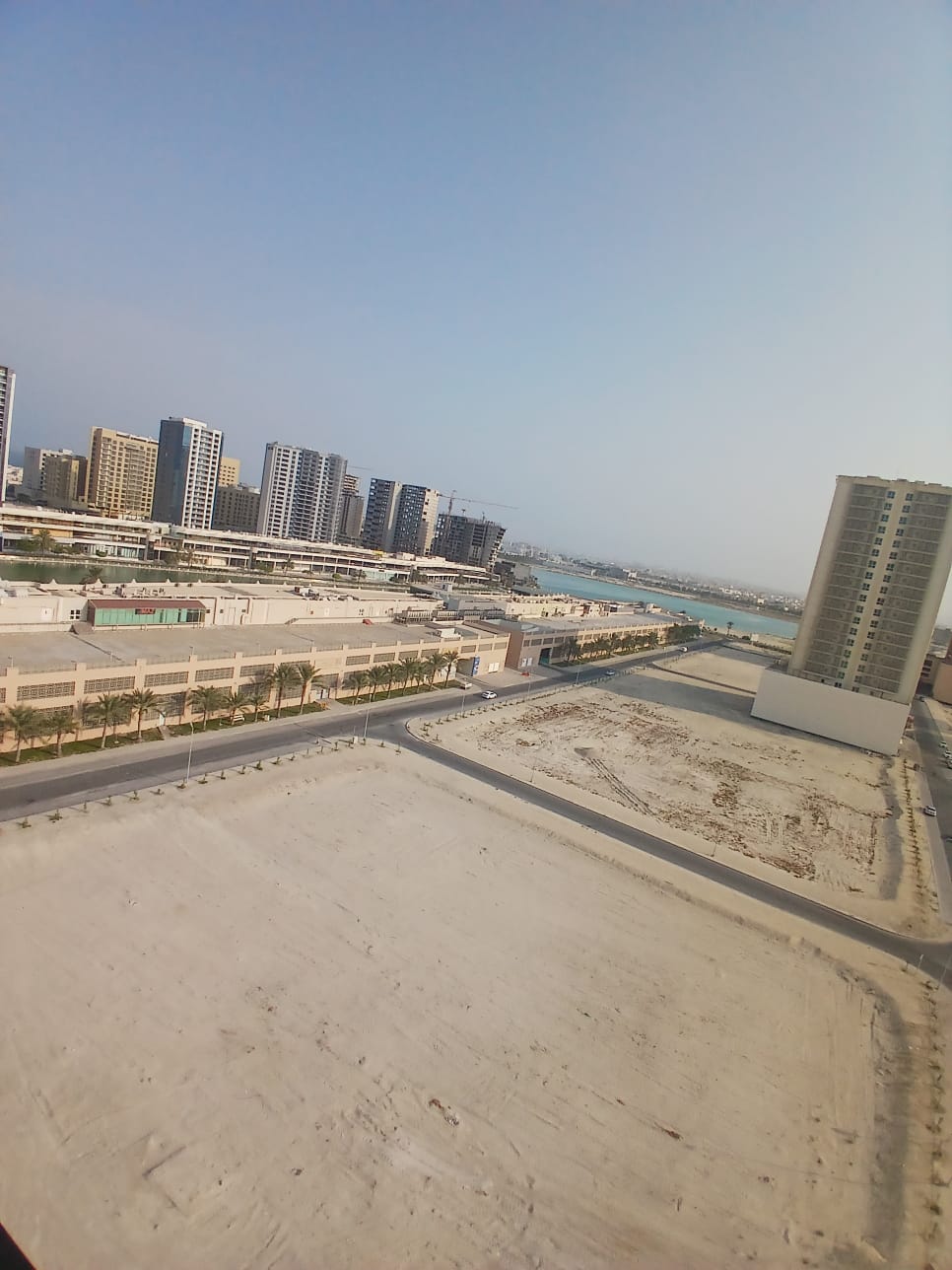 A cityscape view from a high angle showing a collection of modern buildings, a sparsely populated area with sandy plots, and a distant body of water under a clear sky.