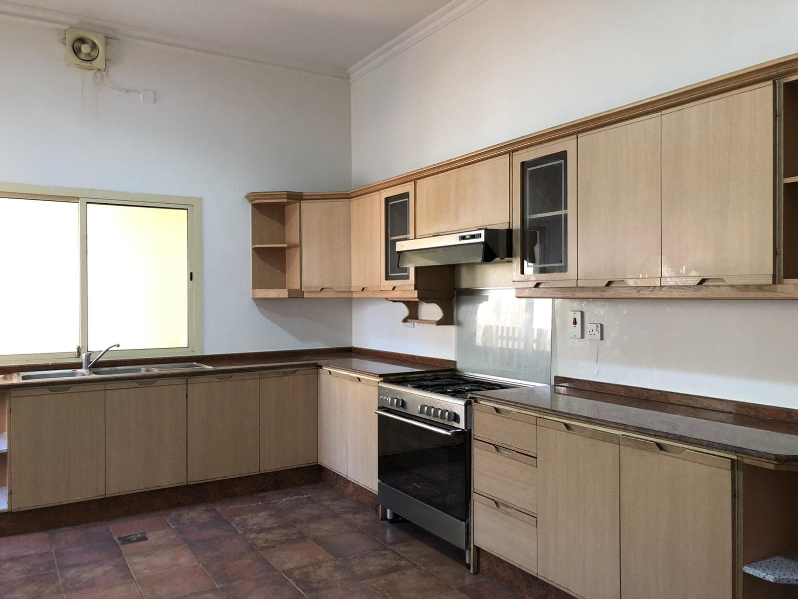 A kitchen with wooden cabinets, a stove with an oven, and a sink under a window. The floor is tiled, and there is an exhaust fan near the ceiling.