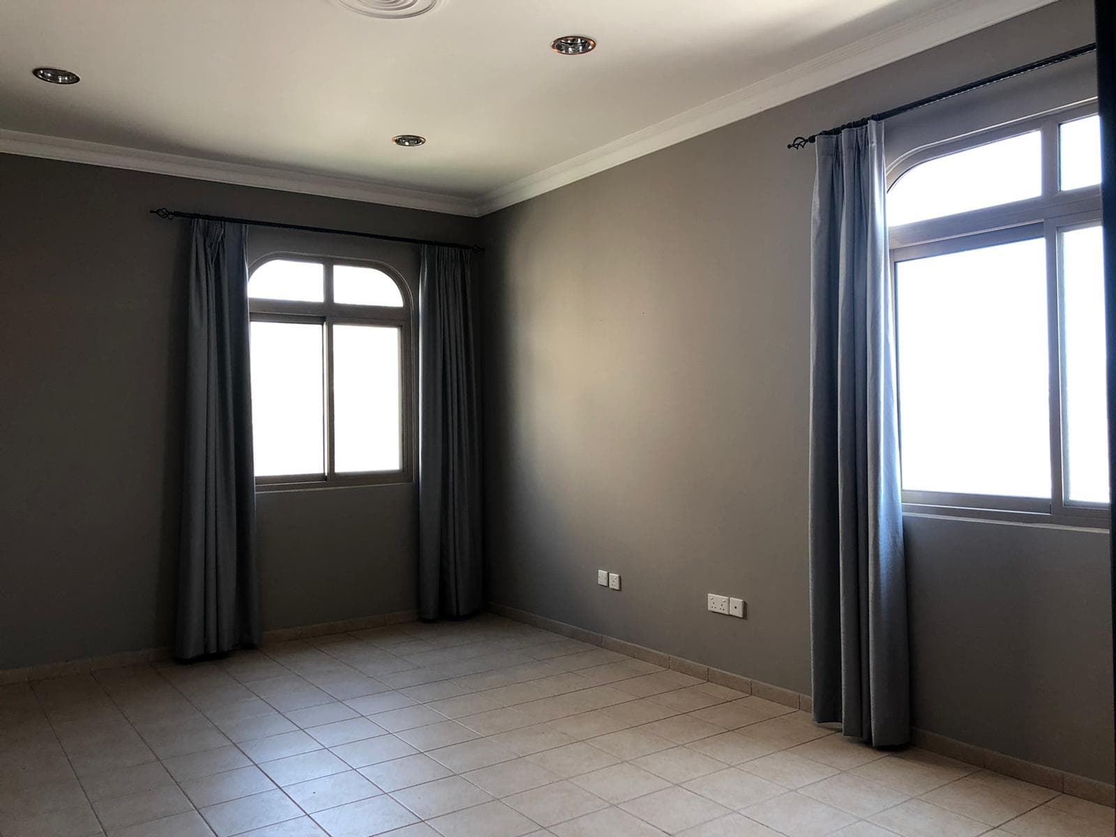 Empty room with gray walls, tile flooring, two windows with gray curtains, and ceiling lights.