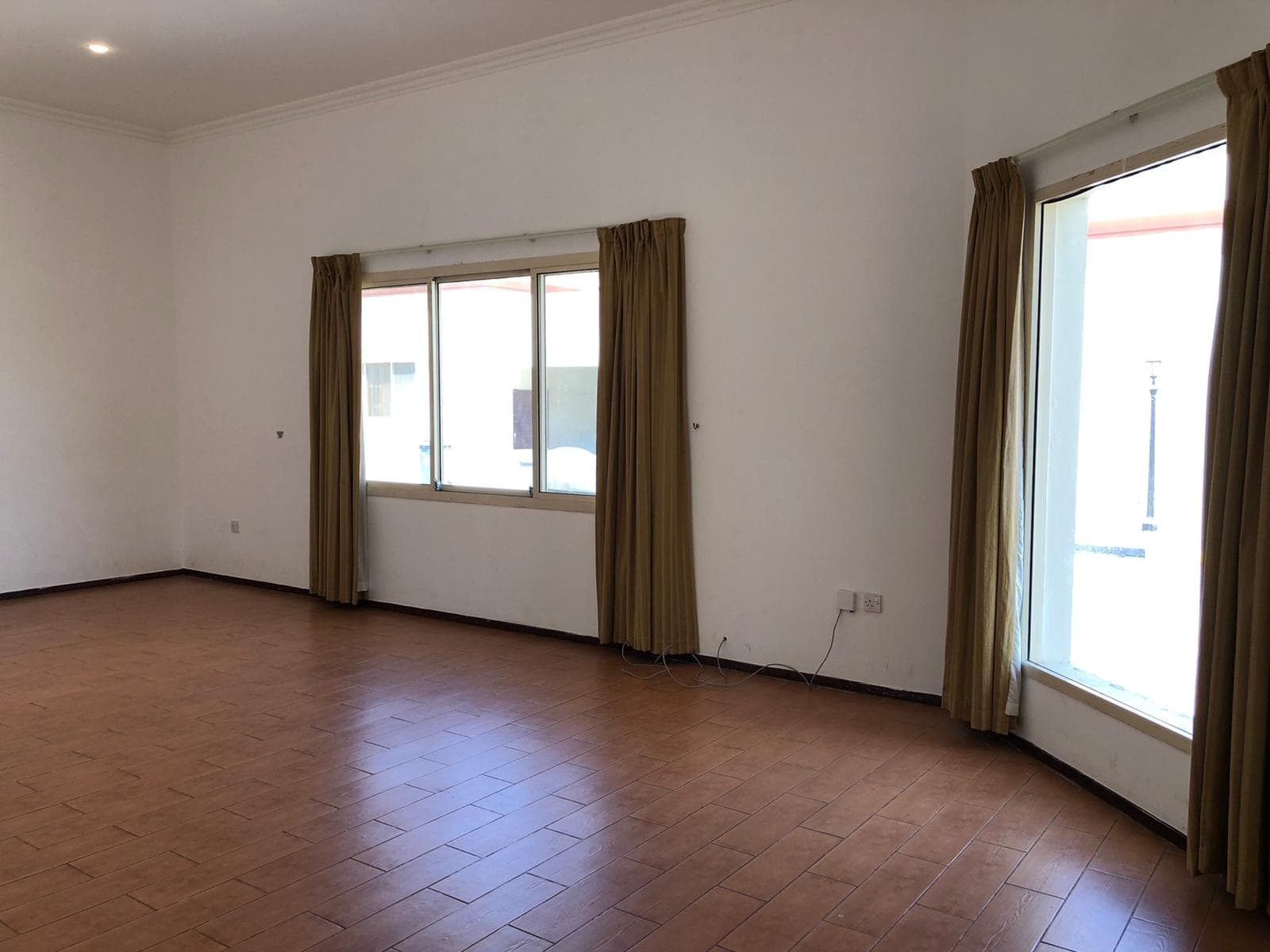 An empty room with tiled flooring, white walls, and mustard-colored curtains on two windows, one on the left and one on the right wall. The room is well-lit with natural light.