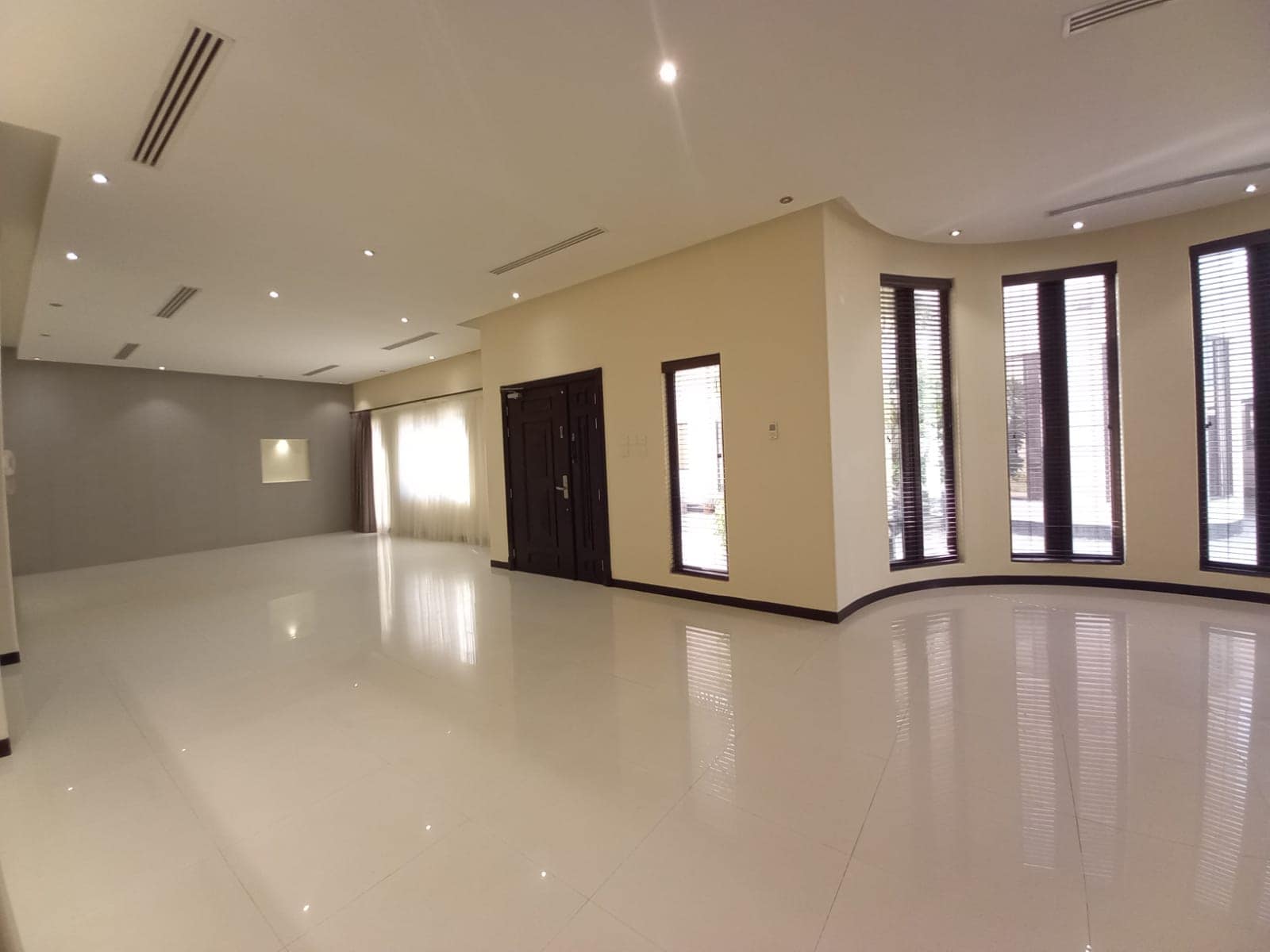 A spacious, unfurnished room with white tiled floor, beige walls, a central dark wooden door, large windows with blinds, and ceiling lights.