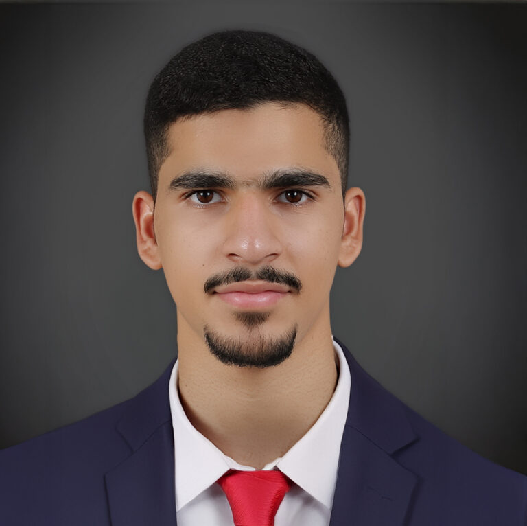A young man with short black hair, trimmed beard, and mustache, wearing a dark suit, white shirt, and red tie, poses against a plain dark background.
