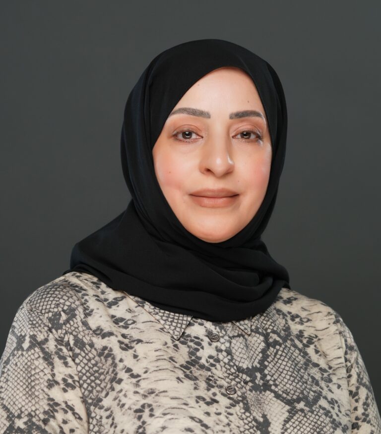 A woman wearing a black hijab and a patterned top poses in front of a plain dark background, looking at the camera with a neutral expression.