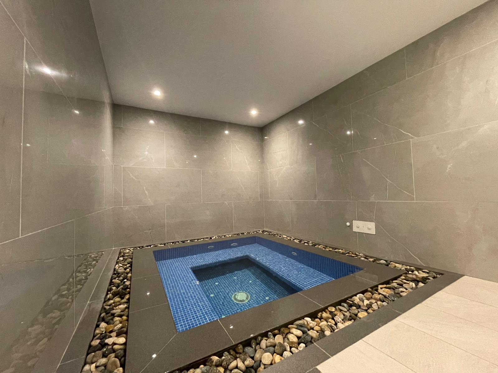 Indoor jacuzzi with blue tile, set in a gray-tiled room, surrounded by decorative stones and lit by recessed ceiling lights.