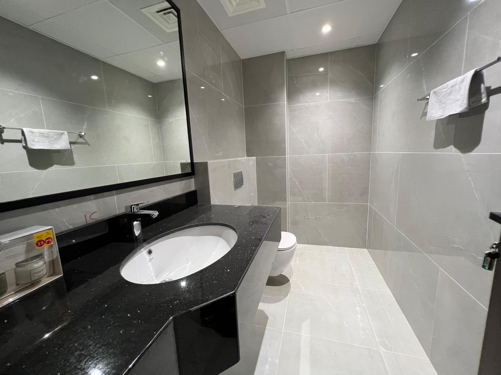 Modern bathroom with gray tiled walls and floor, black countertop with sink, wall-mounted toilet, mirror, towel racks, and toiletries.