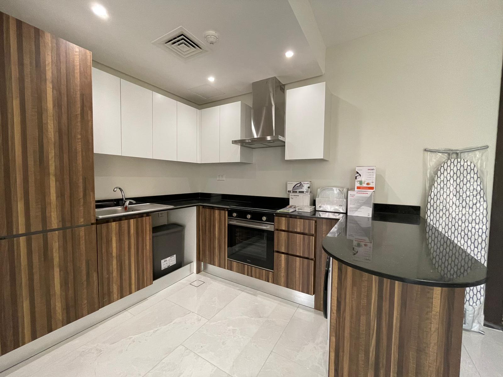 Modern kitchen with wood and white cabinets, black countertops, built-in oven, stovetop, sink, appliances, and an ironing board against a light-colored wall and floor.