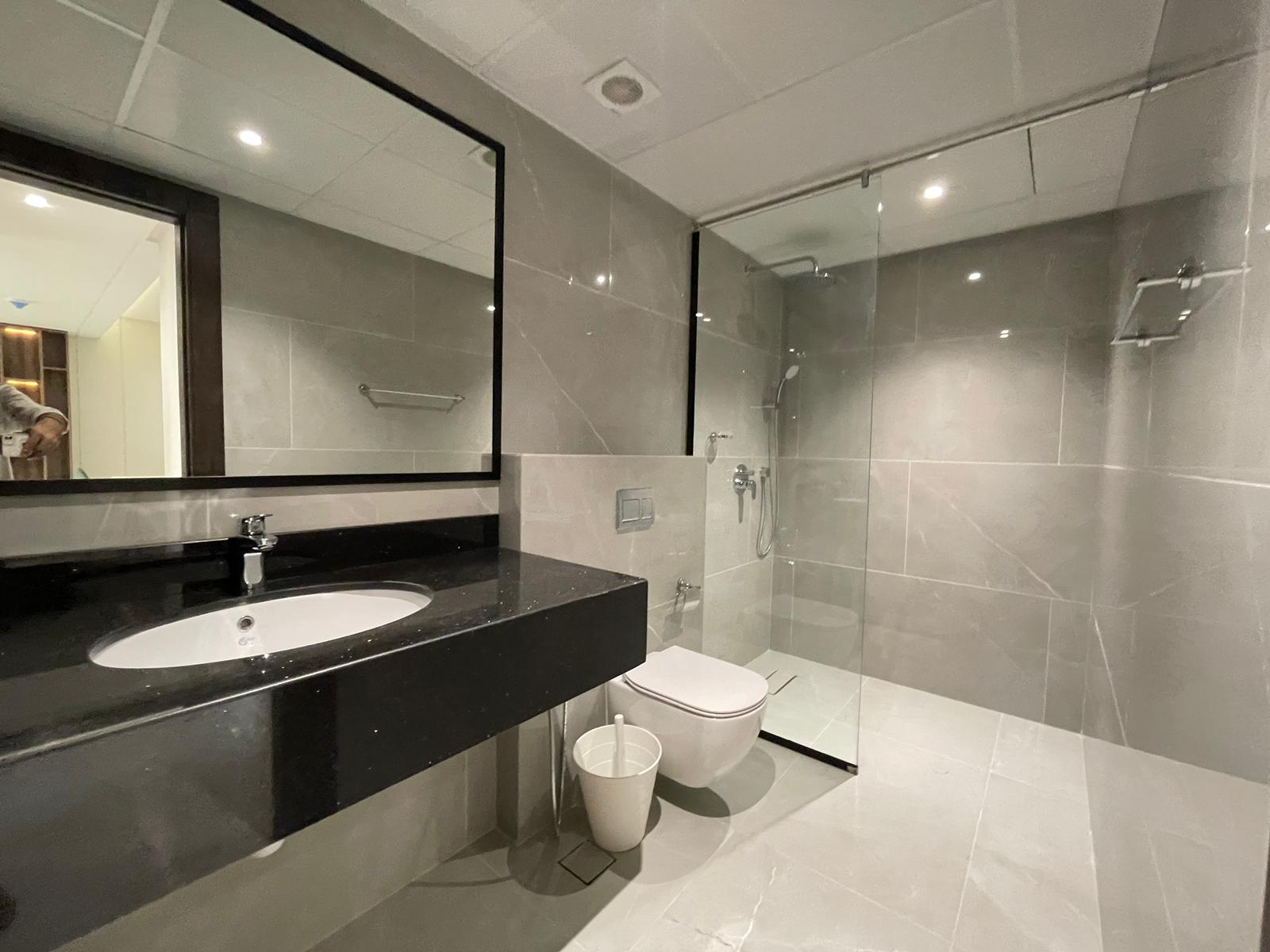 Modern bathroom with a large mirror, black countertop sink, wall-mounted toilet, glass shower enclosure, and beige tiled walls and floor.