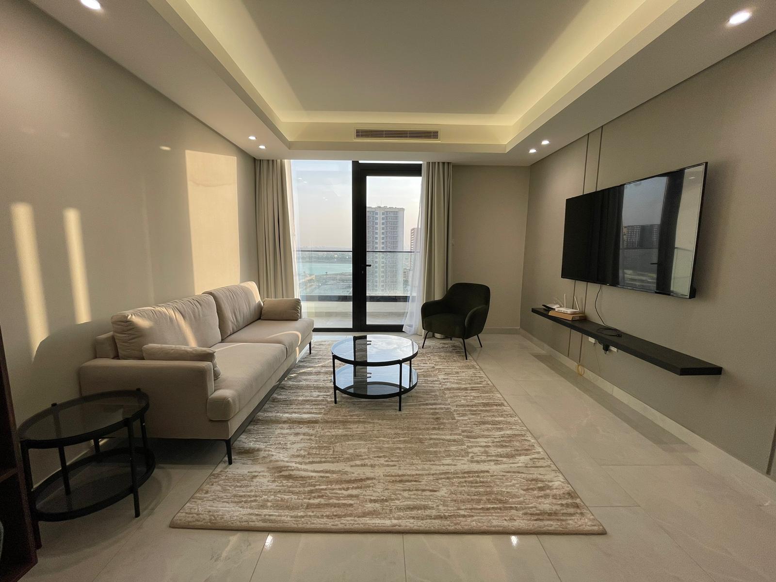 Modern living room with a beige sofa, glass coffee table, black chair, wall-mounted TV, and a large window overlooking city buildings. Natural light fills the space.