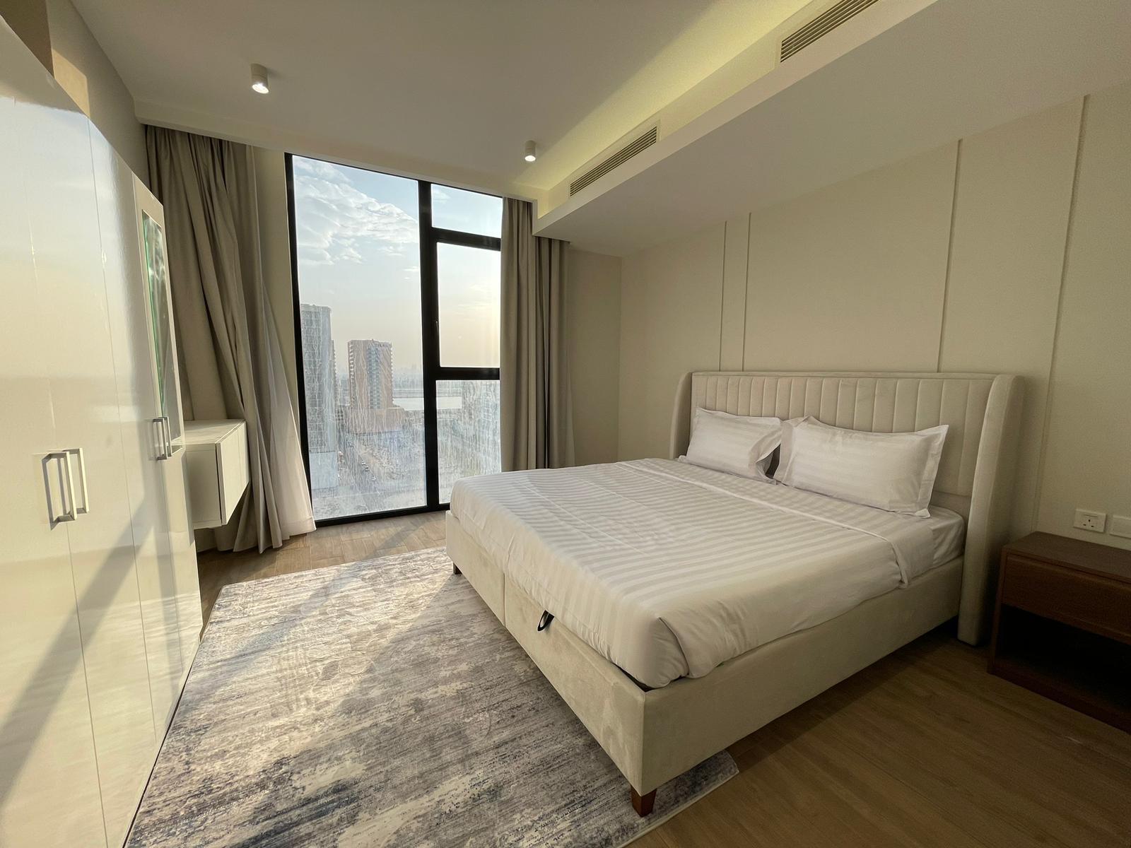 A modern bedroom with a large bed, white linens, a patterned rug, floor-to-ceiling window, and city view in the background.